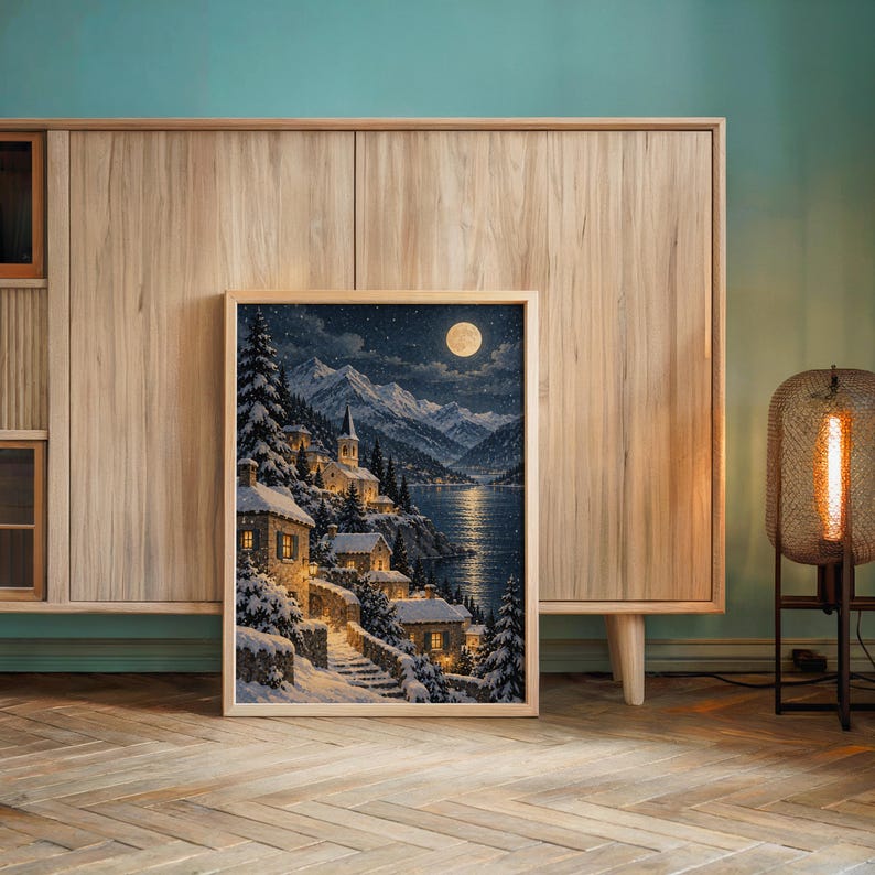 Snowy mountain village wall art print leaning against a wooden cabinet by a teal wall beside a glowing floor lamp.