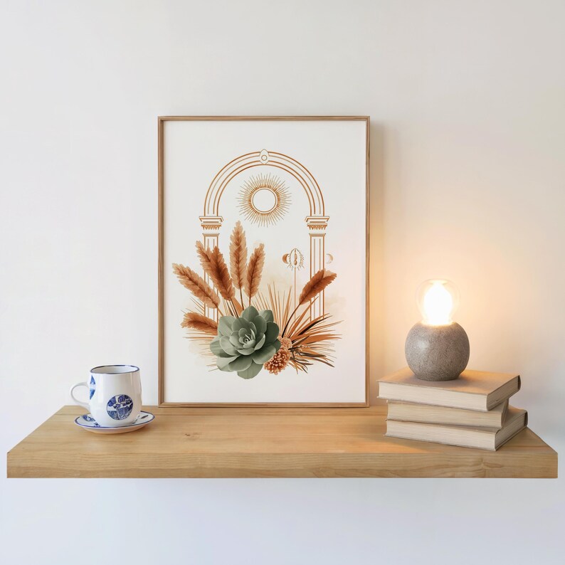 Boho arch wall art featuring a celestial sun, sage green succulent, and pampas grass botanical illustration framed in a light wood frame resting on a floating wooden shelf next to a burning candle, stacked books, and a ceramic teacup in a warm aesthetic interior decor setup.