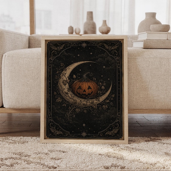 Vintage halloween moon wall art print featuring a burnt orange jack-o-lantern resting on a crescent moon, surrounded by antique gold stars, bats, curling vines, and a deep black celestial sky, framed and leaning against a beige sofa on a textured rug in a bright Scandinavian living room.