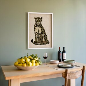A framed print of a sitting leopard with dark green and brown spots on a light background, hanging on a light green wall above a wooden table set with wine and lemons.