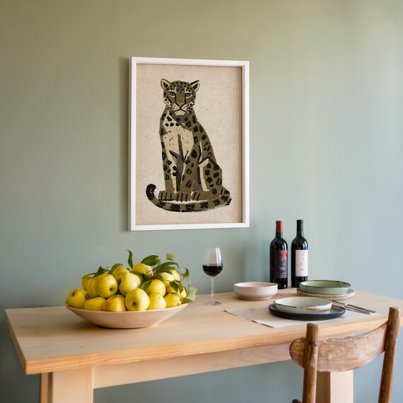 A framed print of a sitting leopard with dark green and brown spots on a light background, hanging on a light green wall above a wooden table set with wine and lemons.