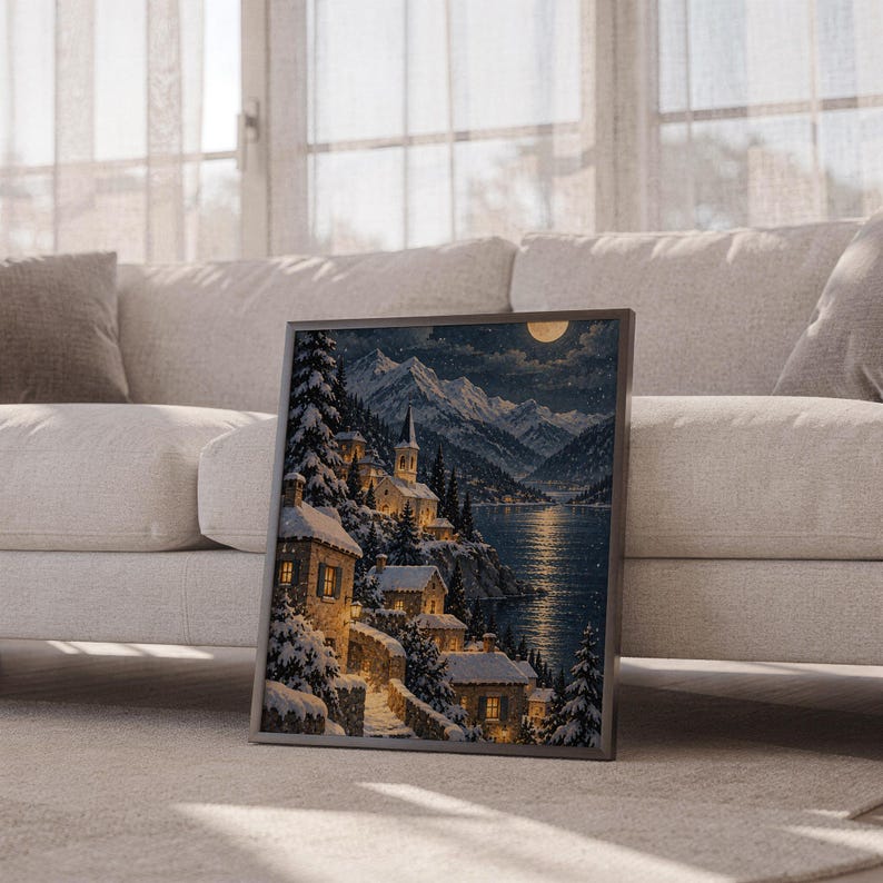 Snowy mountain village wall art print leaning against a white sofa in a bright living room with sheer curtains.