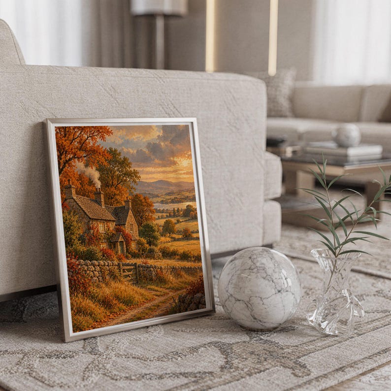 Autumn cottage landscape wall art print in a silver frame leaning against a sofa beside a decorative marble sphere.