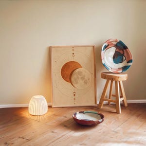 Sun and moon celestial wall art print leaning against a warm beige wall on a wooden floor beside a stool and a glowing lamp.