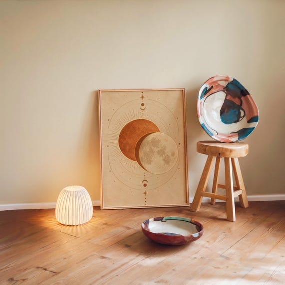 Sun and moon celestial wall art print leaning against a warm beige wall on a wooden floor beside a stool and a glowing lamp.