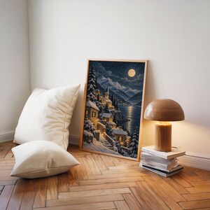 Snowy mountain village wall art print leaning on a herringbone wood floor beside cream cushions and a wooden mushroom lamp.