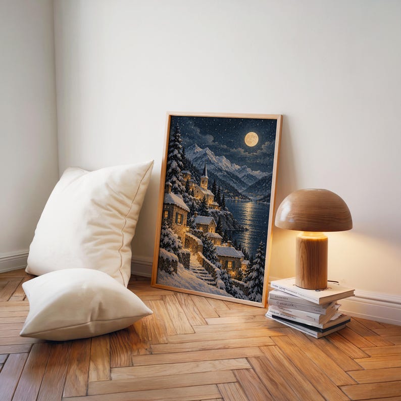 Snowy mountain village wall art print leaning on a herringbone wood floor beside cream cushions and a wooden mushroom lamp.
