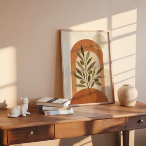 Olive branch art print on dark wooden desk with stacked books and sunlight