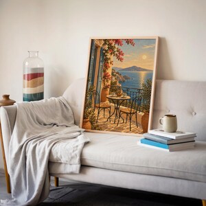 Mediterranean balcony wall art print leaning on a cream sofa against a beige wall beside books and a mug.