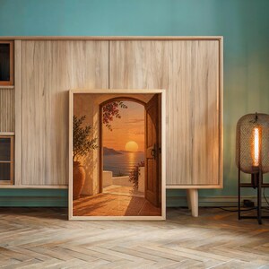 Mediterranean sunset doorway wall art print leaning against a wooden cabinet by a teal wall beside a glowing floor lamp.