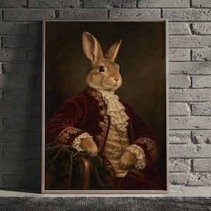 Regal rabbit portrait wall art print featuring a dignified brown rabbit dressed in a deep burgundy velvet coat with ornate gold embroidery and ivory lace, set against a moody dark background in classical Victorian oil painting style, framed and leaning against a beige sofa on a textured rug in a bright Scandinavian living room.