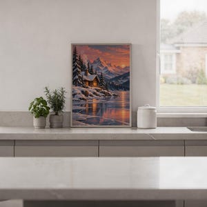 Winter cabin mountain wall art print standing on a white kitchen counter beside potted herbs.