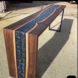 Puede incluir: Una consola larga de madera con un río de resina epoxi azul oscuro en el centro. La mesa tiene un acabado de vetas de madera marrón oscuro y una pata de metal negro. Una pieza de mobiliario única, de unos 200 cm de largo.