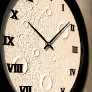 May include: A round wall clock with a textured, off-white face and black Roman numerals. The clock has black hands and a black frame. The face has a unique, cratered appearance, adding a distinctive touch to the design.