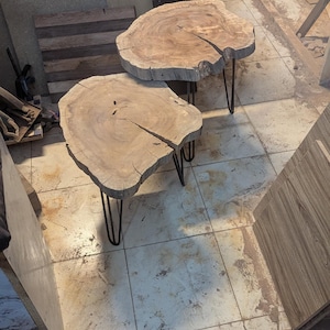 May include: Two handcrafted wooden side tables with natural edges and dark metal hairpin legs. The light brown tabletops have visible wood grain and cracks, showcasing a rustic aesthetic. The tables are approximately 24 inches in diameter.
