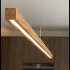Puede incluir: Una lámpara colgante rectangular de madera con una fuente de luz LED lineal y brillante. La luz está suspendida del techo por finos cables. La madera tiene una veta natural y un tono cálido, creando un diseño moderno y minimalista.