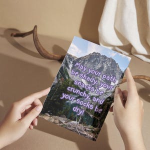 May include: A rectangular card with a mountain landscape image and the text "May your path be shady, your snacks be crunchy, and your socks be dry!" The card is held by hands against a neutral background.
