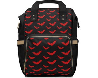 May include: Black backpack with a repeating pattern of red bats. The bag has a top handle, a front pocket, and side pockets. The zippers and trim are gold-colored.
