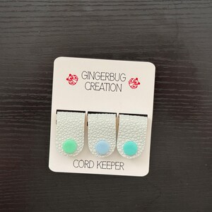 May include: Three cord keepers on a white card. Each cord keeper is white with a textured surface and a colored circle at the bottom. The colors are green, blue, and green. The card reads "GINGERBUG CREATION" and "CORD KEEPER".