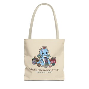 May include: A cream-colored tote bag featuring a graphic of a blue dragon knitting with yarn. The illustration includes a basket, a container of supplies, and a pincushion. Below the graphic, the text reads Stitch's Patchwork Cottage Made with Heart.