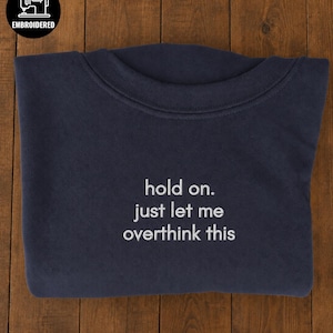 Pode incluir: Uma camisola azul-marinho de gola redonda com texto bordado a branco que diz: "hold on. just let me overthink this." A peça tem uma gola canelada e mangas compridas, com um logótipo preto circular no canto que diz "EMBROIDERED."