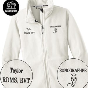 Könnte beinhalten: Cremefarbene Fleecejacke mit durchgehendem Reißverschluss und Stehkragen. Die Jacke trägt die gestickten Schriftzüge "Taylor RDMS, RVT" und "SONOGRAPHER" mit einer Ultraschallgrafik. Ein schwarz-weiß gesticktes Nähmaschinenlogo befindet sich oben links.