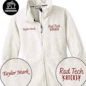 May include: A white fleece jacket with a full zipper and a high collar. The jacket has the embroidered text "Taylor Mark" on the left chest and "Rad Tech RT(R)" on the right chest in red. A black and white embroidered patch that says "EMBROIDERED" is in the upper left corner.