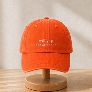 May include: A bright orange baseball cap featuring the white embroidered text will yap about books centered on the front panel. The cap has a structured crown, a curved brim, and a button at the top, showcasing a classic six-panel design.