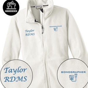 Könnte beinhalten: Eine weiße Fleecejacke mit durchgehendem Reißverschluss und Stehkragen. Auf der linken Brust ist der Name "Taylor RDMS" blau gestickt, und auf der rechten Brust steht das Wort "SONOGRAPHER" mit einer Sonogramm-Grafik. Ein "EMBROIDERED" Logo ist ebenfalls vorhanden.