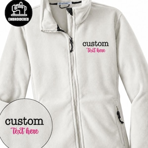 Puede incluir: Una chaqueta de forro polar blanca con cremallera frontal completa, cuello alto y dos bolsillos laterales con tiradores. El pecho presenta el texto bordado custom Text here en negro y rosa, junto con un detalle circular en la imagen.