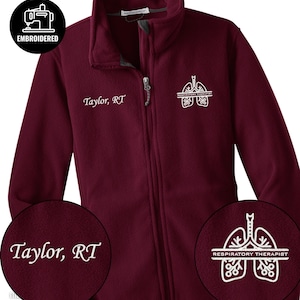Puede incluir: Chaqueta polar granate con cremallera completa y cuello alto. La chaqueta tiene el nombre "Taylor, RT" bordado en el pecho y un gráfico blanco de pulmones con las palabras "Respiratory Therapist". Un logotipo circular indica que la chaqueta está bordada.