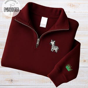 May include: A maroon quarter-zip sweatshirt with a silver zipper and a small embroidered zebra on the chest. The sleeve features a green embroidered design. The words "Personalize it" and "Embroidered" are printed on the top left.