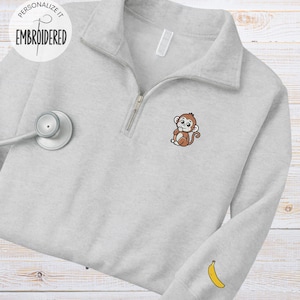 Puede incluir: Sudadera gris claro con cremallera de cuarto de cuello con un pequeño mono bordado en el pecho y un plátano en la manga. El texto "PERSONALIZE IT EMBROIDERED" está en un círculo en la parte superior izquierda.
