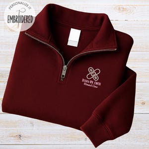 May include: A maroon quarter-zip sweatshirt with a silver zipper. The sweatshirt has the text "Personalize it Embroidered" in the upper left corner. The lower right side of the sweatshirt has a white graphic of two bandages crossed with the text "Jessica RN, CWCN Wound Care".