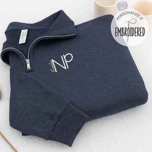 May include: A navy blue quarter-zip sweatshirt with a silver zipper and embroidered initials "NP". The sweatshirt has a ribbed cuff and collar. The image includes the text "Personalize it" and "Embroidered".