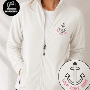 Puede incluir: Una chaqueta polar de color crema con cremallera y un diseño de ancla bordado en negro en el pecho izquierdo. Debajo del ancla, el texto YOUR BOAT NAME aparece en rosa. La chaqueta tiene cuello alto, mangas largas y cierre de cremallera.