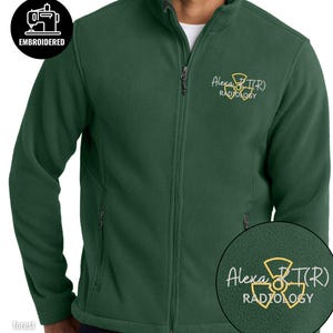 May include: Forest green fleece jacket with a full zipper and a stand-up collar. The jacket features an embroidered logo that says "Alexa R(T)(R) Radiology" in yellow. The jacket also has two side pockets.