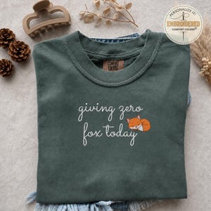 May include: A dark green t-shirt featuring white cursive text that reads giving zero fox today, accompanied by an embroidered graphic of a small, sleeping orange fox. The shirt has a crew neckline and a visible brand tag on the inner collar.