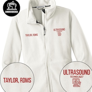 Puede incluir: Chaqueta polar blanca con cremallera completa y cuello alto. La chaqueta tiene el texto "TAYLOR, RDMS" bordado en el pecho izquierdo y "ULTRASOUND TECHNOLOGIST" con un gráfico en el derecho. Un parche bordado en blanco y negro está en la esquina superior izquierda.