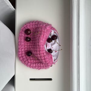 Könnte beinhalten: Ein rosa gestrickter Barett mit strukturiertem Muster, verziert mit mehreren frischen Kirschen. Das Innere des Baretts zeigt einen weißen Stoff mit Kirschmuster. Der Barett liegt auf einer weißen Oberfläche.
