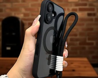 May include: Black phone case with a clear back, featuring a black lanyard with white accents. The case has a magnetic ring and a black border. The phone case is held in a person's hand.