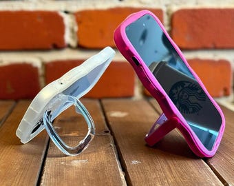 May include: Two phone cases on a wooden surface. One is white with a clear ring holder, the other is hot pink and propped up with a kickstand. The phone screens are visible, reflecting the surroundings.