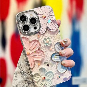 May include: A light pink phone case with a floral design. The case features raised, textured flowers in pink, blue, and white, embellished with small, colourful rhinestones. The phone case has a clear border around the camera.