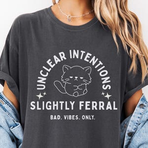 以下が含まれることがあります： 「UNCLEAR INTENTIONS SLIGHTLY FERRAL BAD VIBES. ONLY.」の白文字と猫のイラストがプリントされた濃いグレーのTシャツ。デニムジャケットと合わせて着用されています。