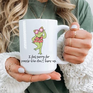 May include: White ceramic mug with a cartoon frog wearing sunglasses and a swimsuit. The frog is green with a red and white polka dot swimsuit. The mug has the text "I feel sorry for people who don't know us."
