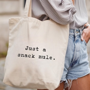 May include: Cream-colored canvas tote bag with the text "Just a snack mule" printed in black. The bag has a simple design and is carried over the shoulder. The bag is a casual accessory.