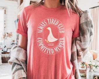 Op de afbeelding: Een koraalkleurig t-shirt met een witte afbeelding van een gans in een cirkelvormig ontwerp. De tekst luidt "ALWAYS TIRED CLUB, COMMITTED MEMBER, COFFEE PLEASE". Het shirt heeft korte mouwen en een ronde hals.