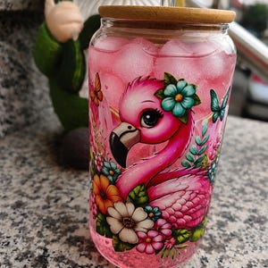 Könnte beinhalten: Ein durchsichtiges Glas mit Bambusdeckel und Glasstrohhalm, gefüllt mit einer rosa Flüssigkeit und Eiswürfeln. Die Außenseite zeigt einen farbenfrohen Grafikdruck eines rosa Flamingos, umgeben von verschiedenen Blumen und Schmetterlingen.