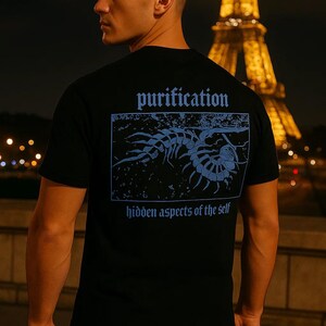 Puede incluir: Camiseta negra con un gr&aacute;fico azul y texto en la espalda. El texto dice "purification" encima de una ilustraci&oacute;n azul de una criatura, con "hidden aspects of the self" debajo. La Torre Eiffel est&aacute; al fondo.