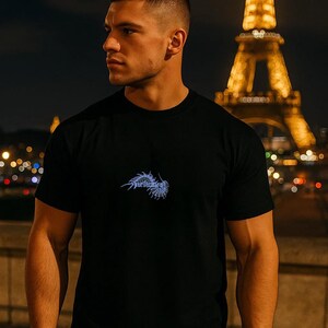 Puede incluir: Camiseta negra de cuello redondo con un dise&ntilde;o gr&aacute;fico azul en el pecho. La Torre Eiffel es visible al fondo, iluminada por la noche. La palabra "Purification" forma parte del gr&aacute;fico.